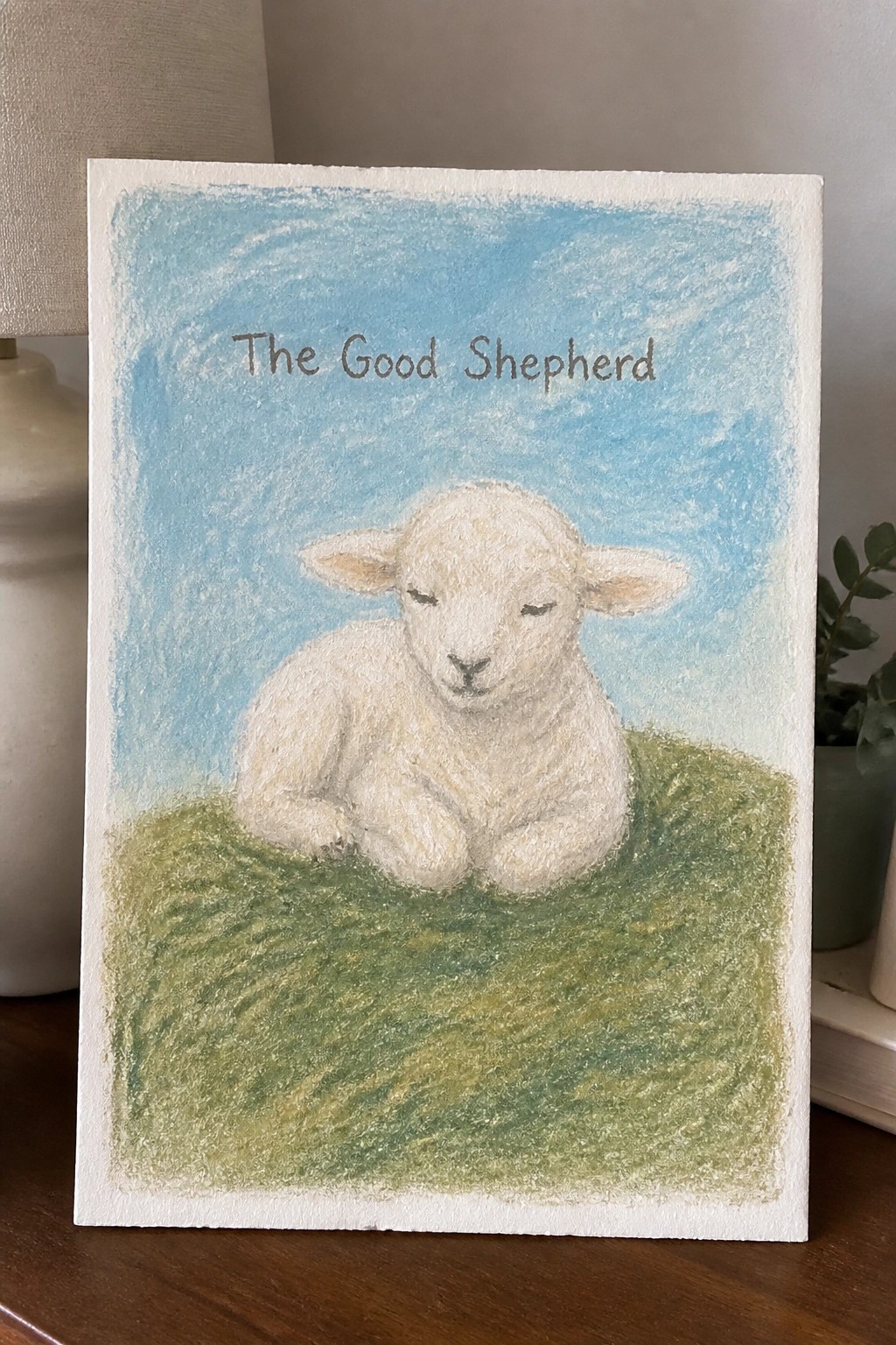 Pastel artwork of white lamb resting on green grass under blue sky titled The Good Shepherd