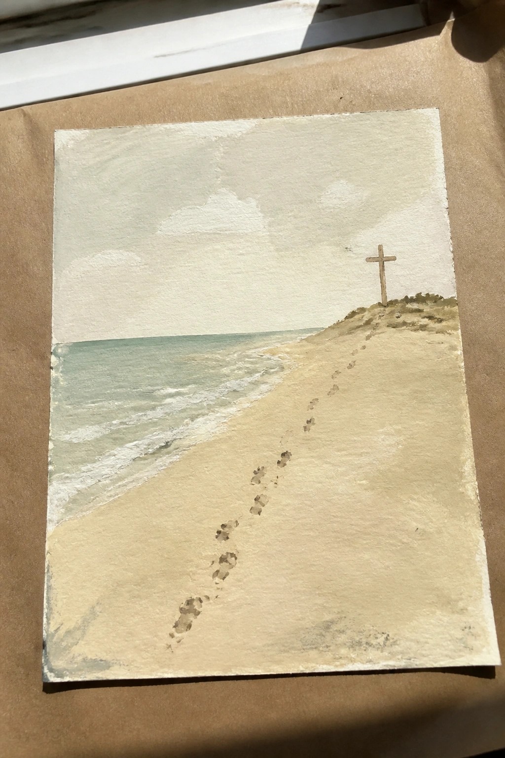 Watercolor painting of beach with footprints in sand leading to wooden cross on dune