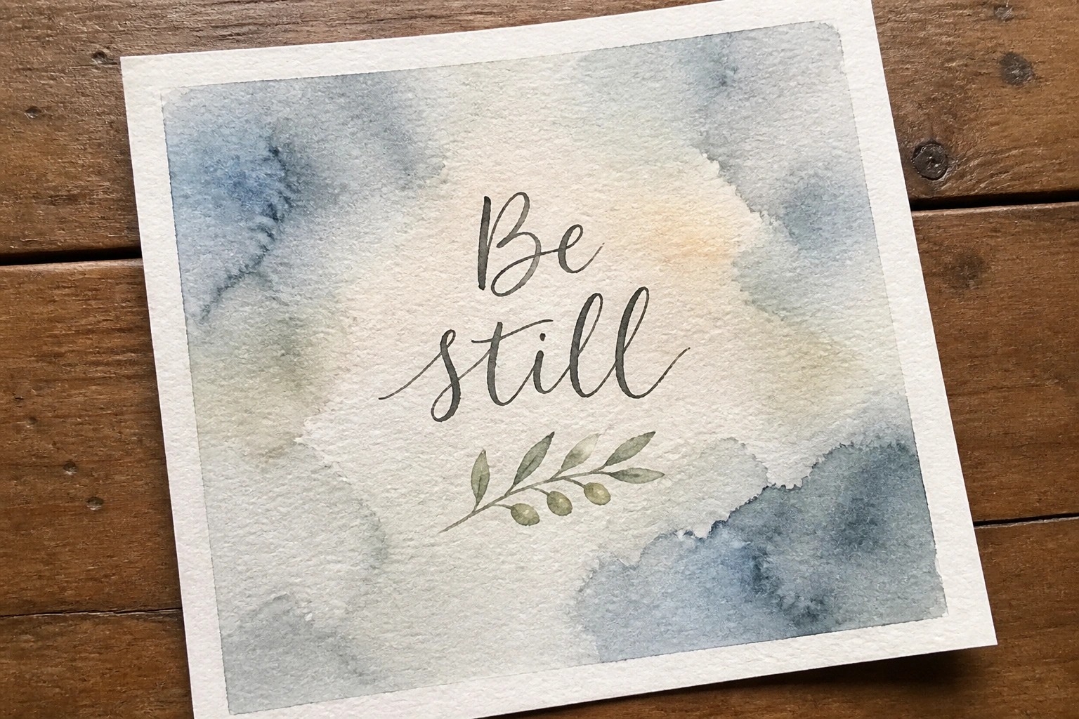 A watercolor painting with the words Be still in black script above a small olive branch on a blue and beige background.
