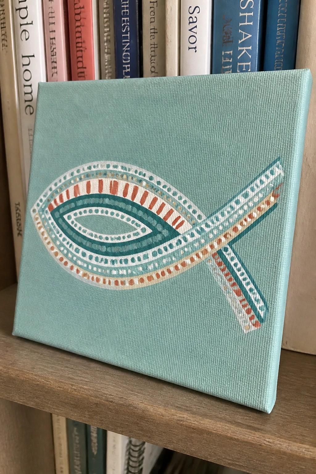 Light teal canvas with ornate patterned ichthys fish on wooden bookshelf among books