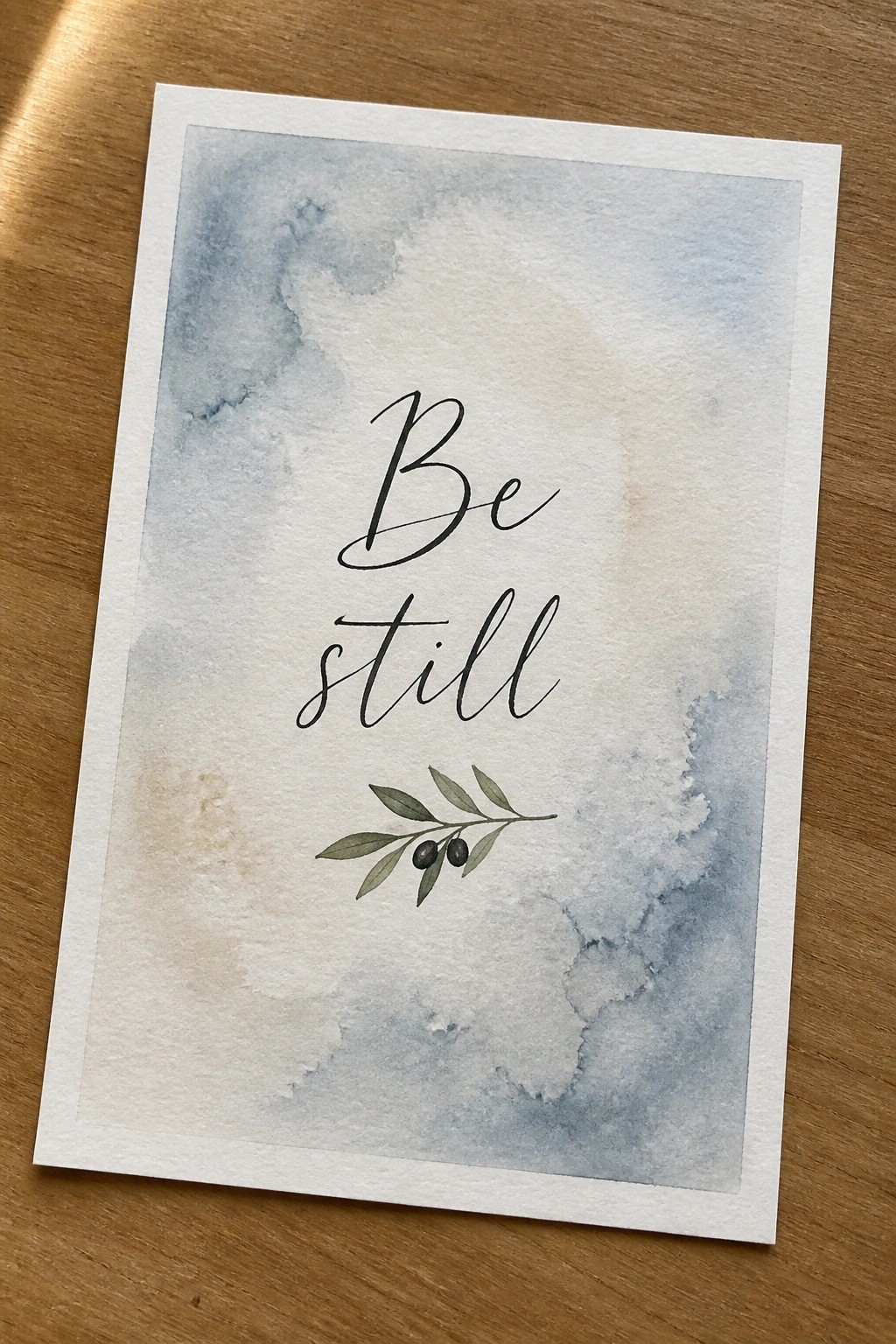 A watercolor painting with the words Be still in black script above a small olive branch on a blue and beige background.