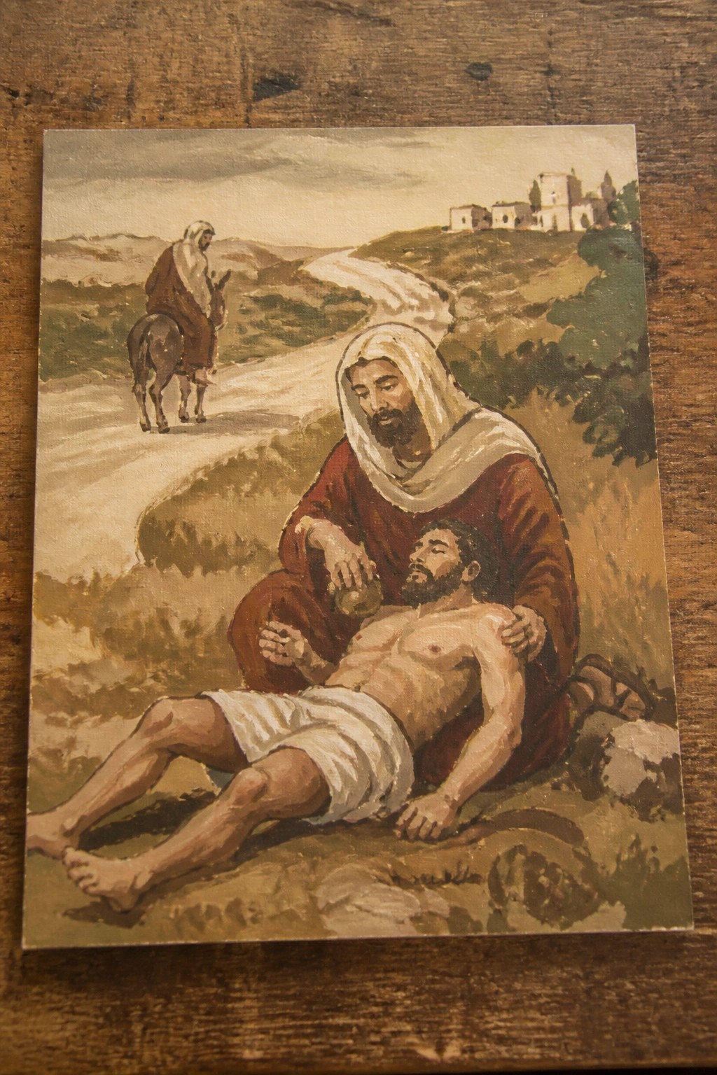 Oil painting of robed man pouring from flask onto shirtless injured figure by path with distant rider