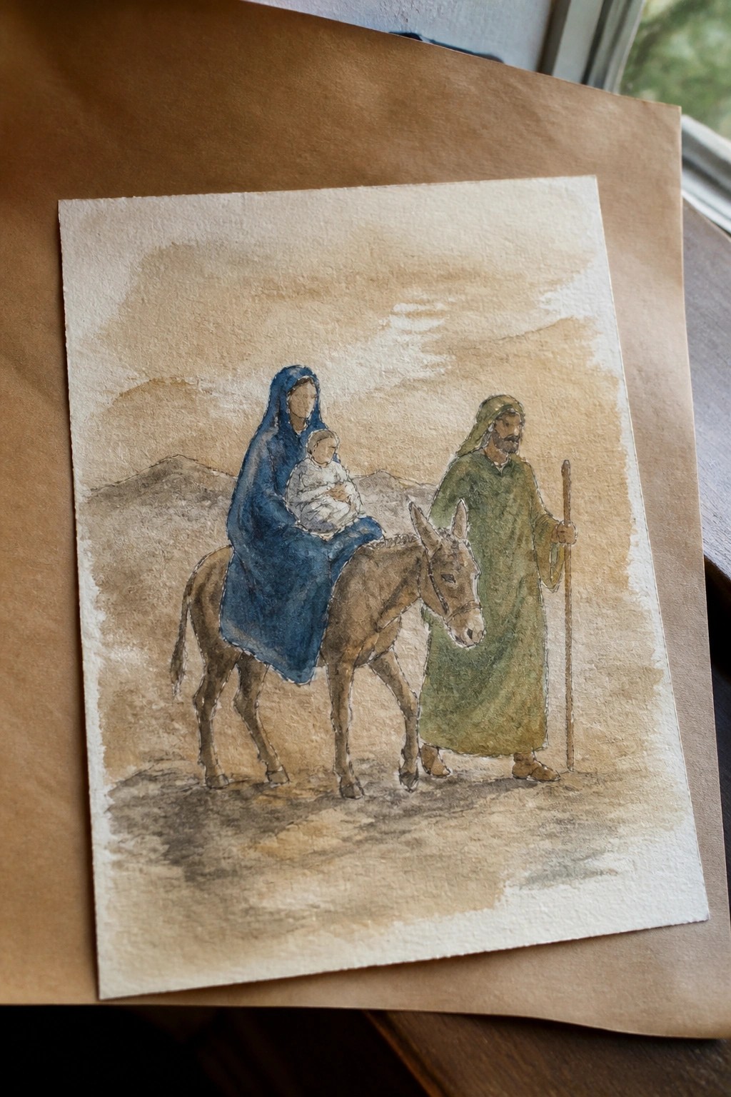 Watercolor of blue-cloaked woman on donkey holding baby beside green-robed man with staff.