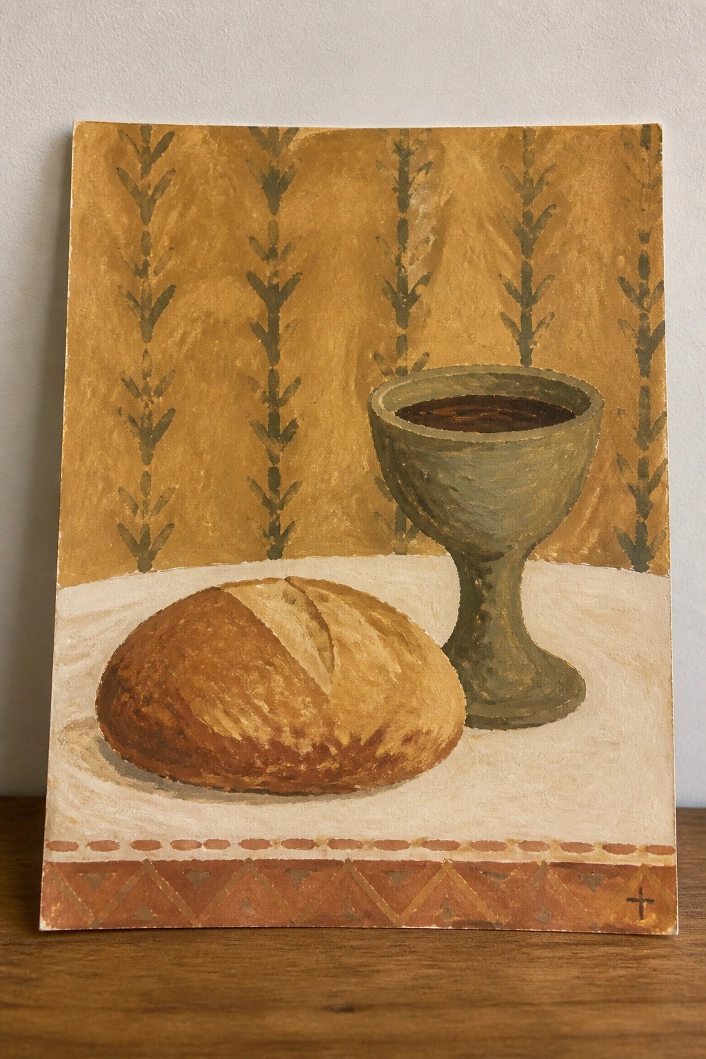 A painting shows a loaf of bread and a green chalice on a white surface against a golden background with leaf patterns and a small cross.