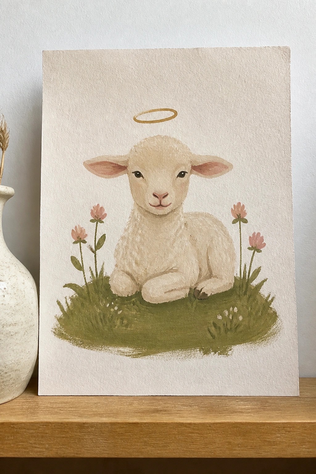 Watercolor of a white lamb with golden halo resting on green grass amid pink flowers.