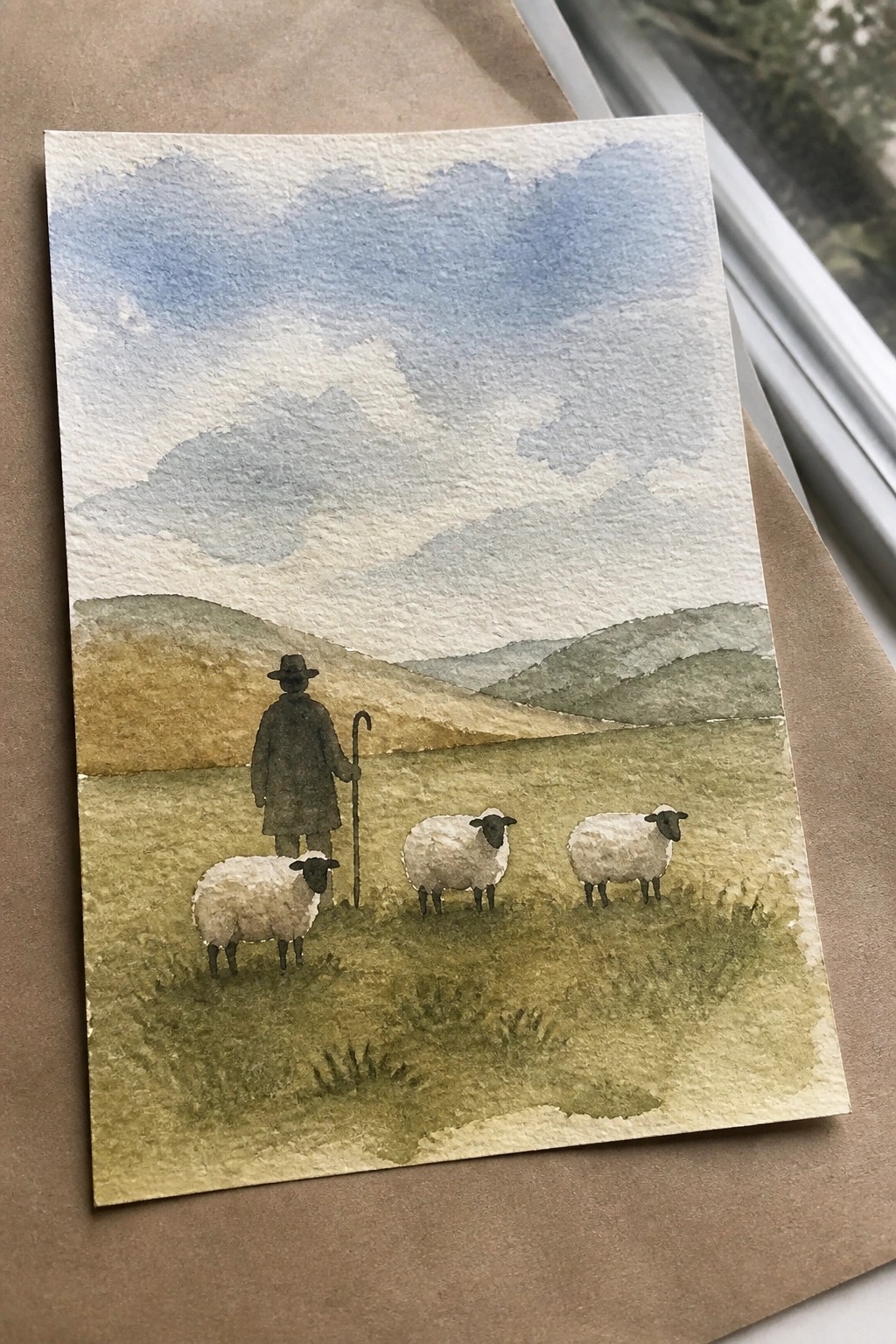 Watercolor of shepherd in dark coat with crook beside three sheep in green hills