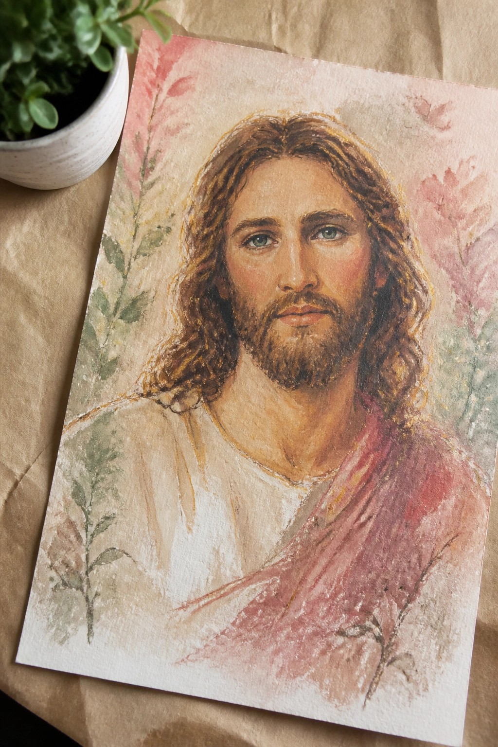 Watercolor portrait of Jesus with long brown hair, beard, white and red robes amid pink flowers.