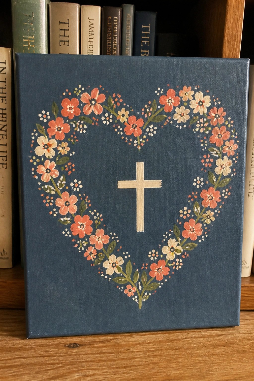 Blue canvas with floral heart wreath encircling white cross on bookshelf