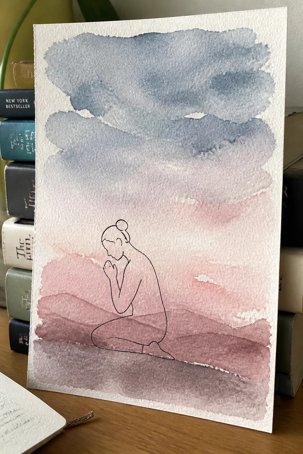 Watercolor of nude kneeling figure over layered blue and pink landscape with books in background.