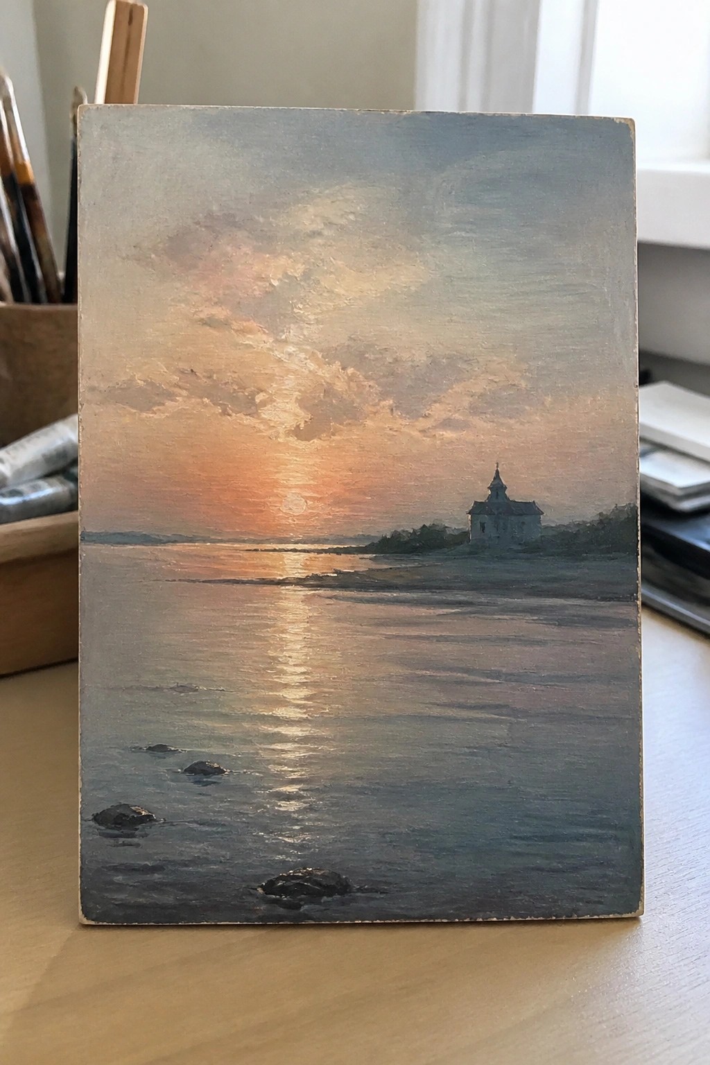 Oil painting of sunset reflecting on calm water with church on shore and rocks.