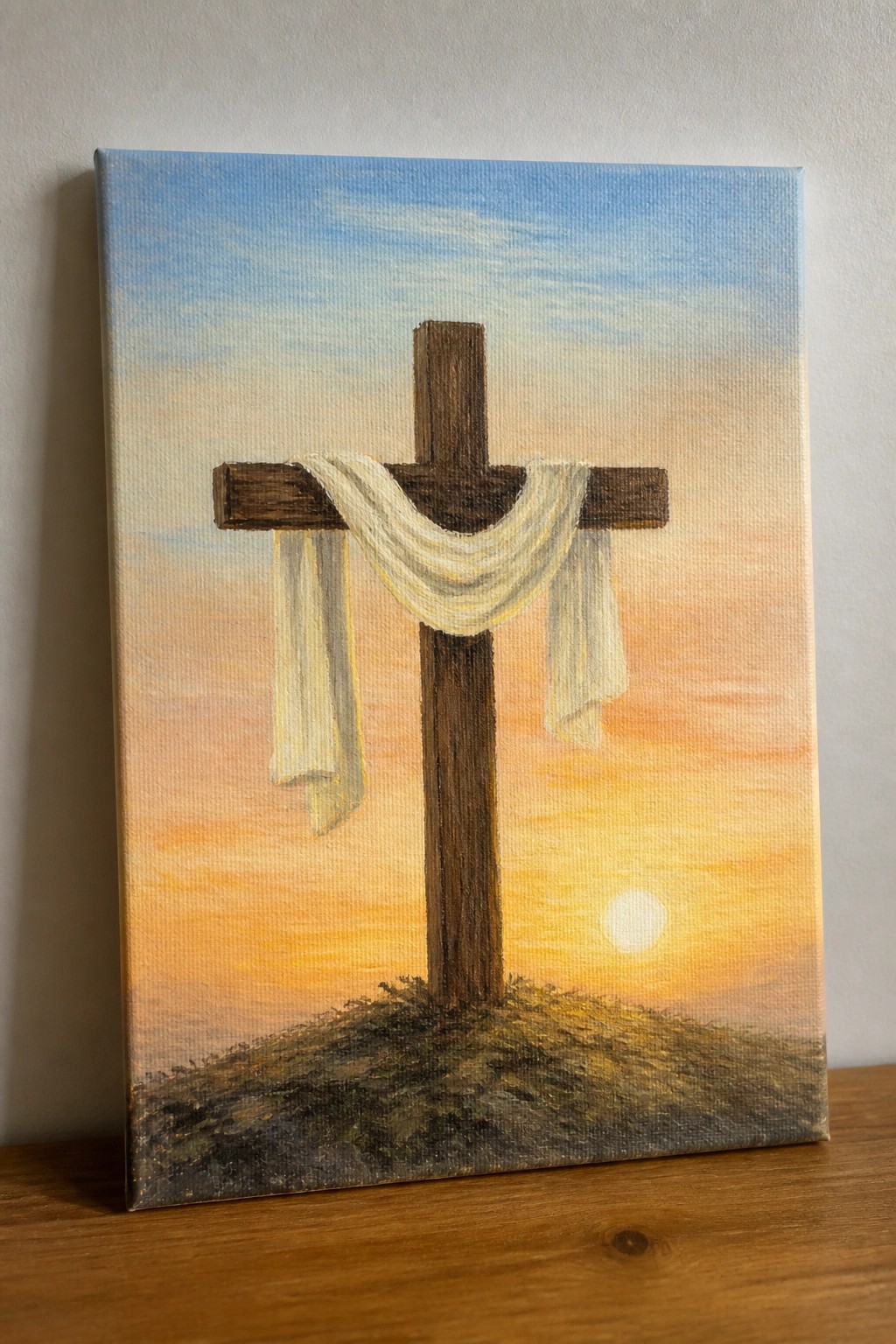 Wooden cross with white cloth on grassy hill under orange sunset sky