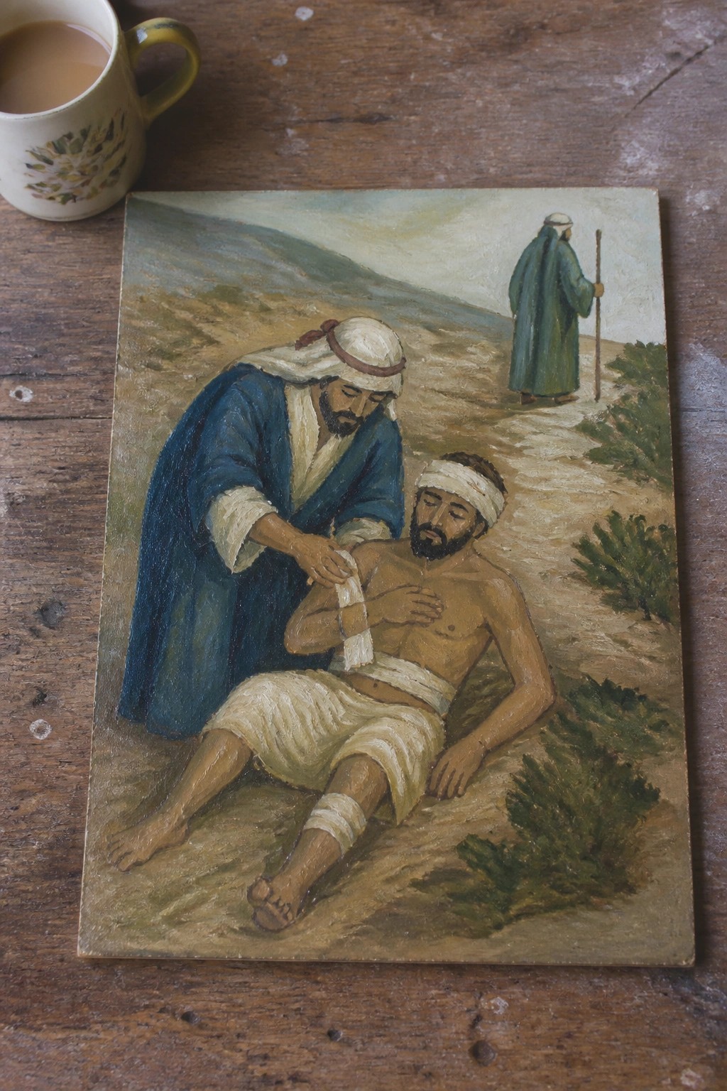 Painting of robed Samaritan bandaging wounded man on path beside floral mug on wood.