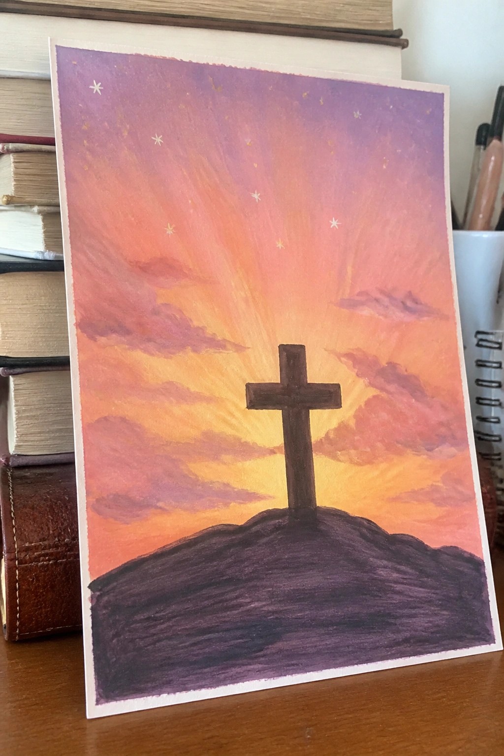 Wooden cross atop dark hill under vibrant purple-orange sunset sky with stars