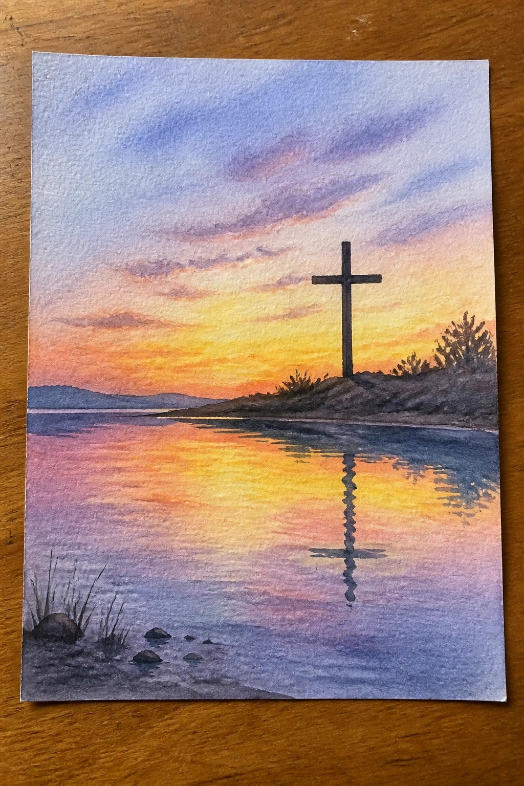 Watercolor painting of silhouetted cross reflected in water beneath vibrant sunset sky.