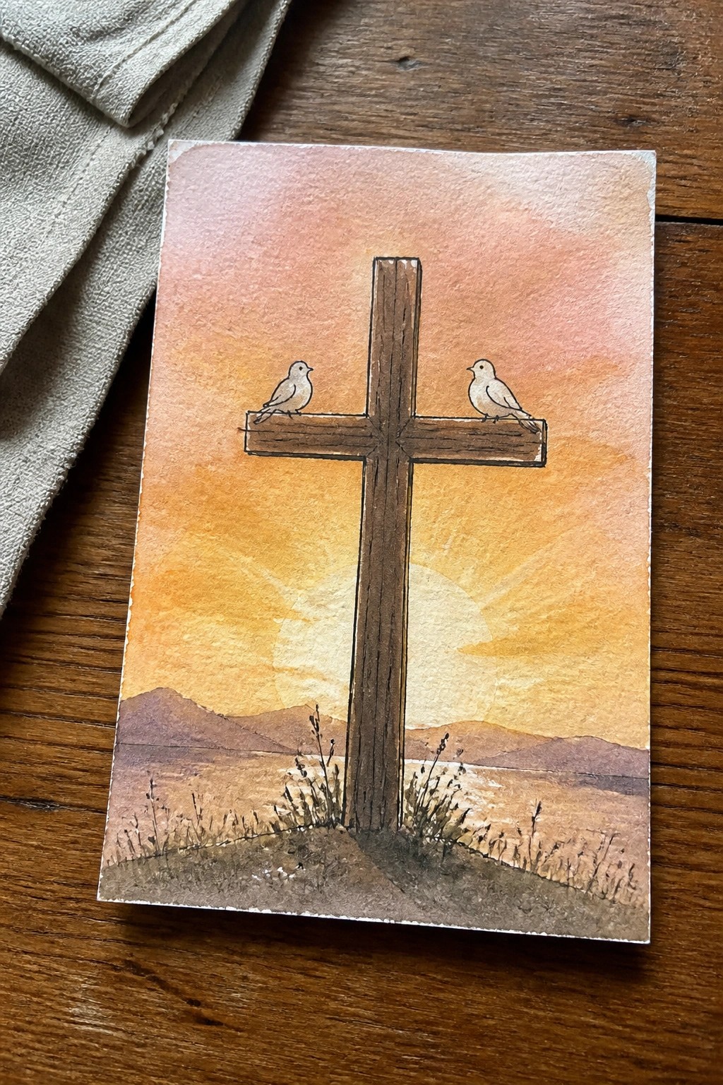 Watercolor cross with two birds perched against orange sunset and distant mountains