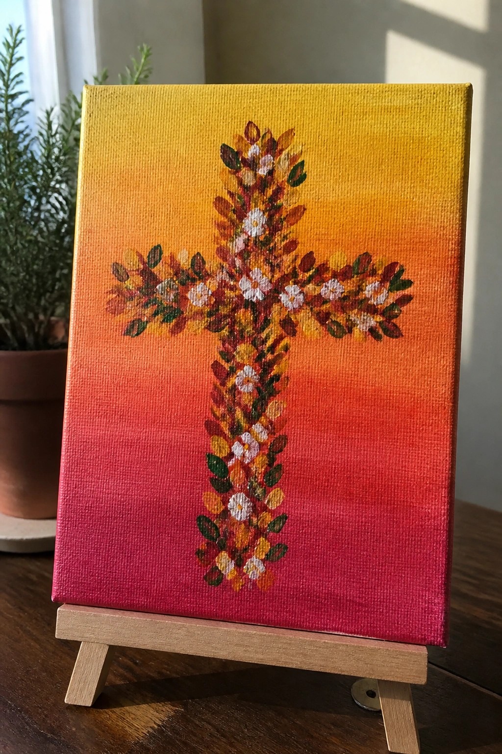 A canvas painting shows a cross made of yellow, orange, red, and green leaves with small white flowers on a yellow-to-red gradient background, placed on a wooden easel.