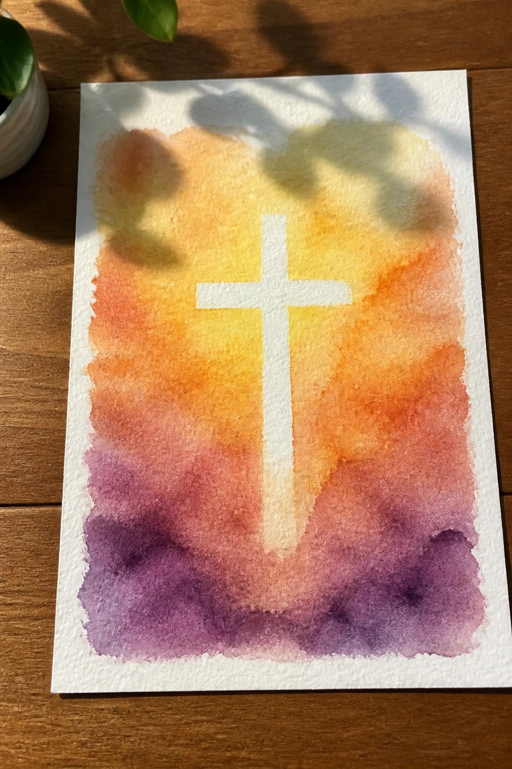 White cross centered on orange-purple watercolor gradient with leaf shadows on wood