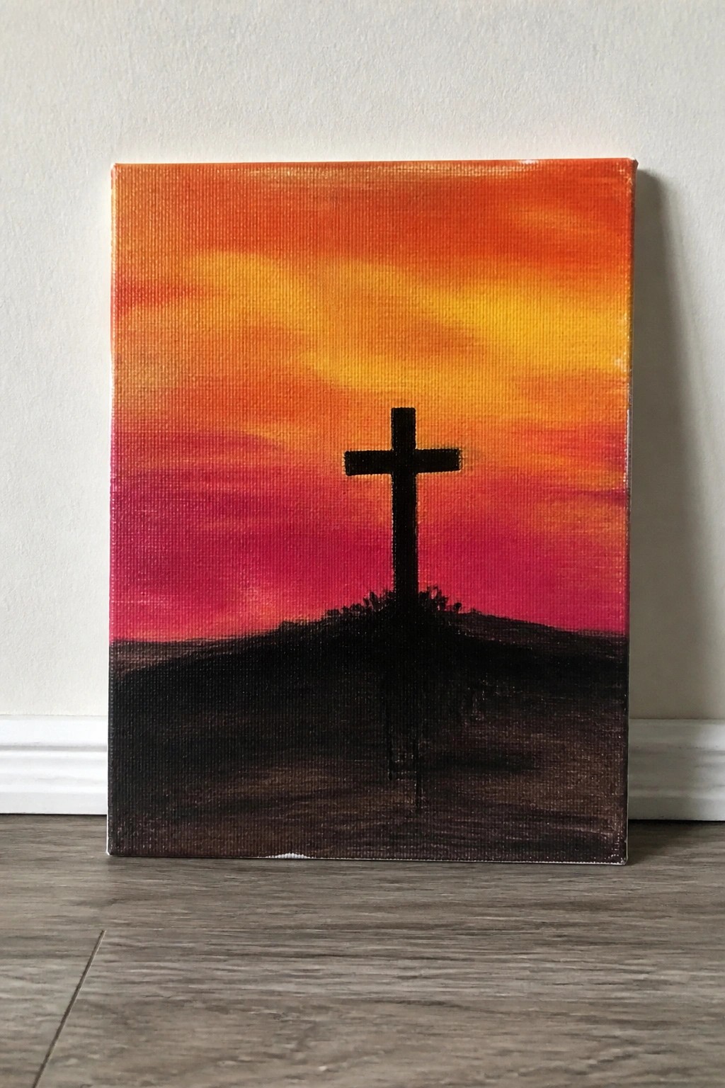 Black cross silhouette on dark hill under vibrant orange-yellow-red sunset sky on canvas.