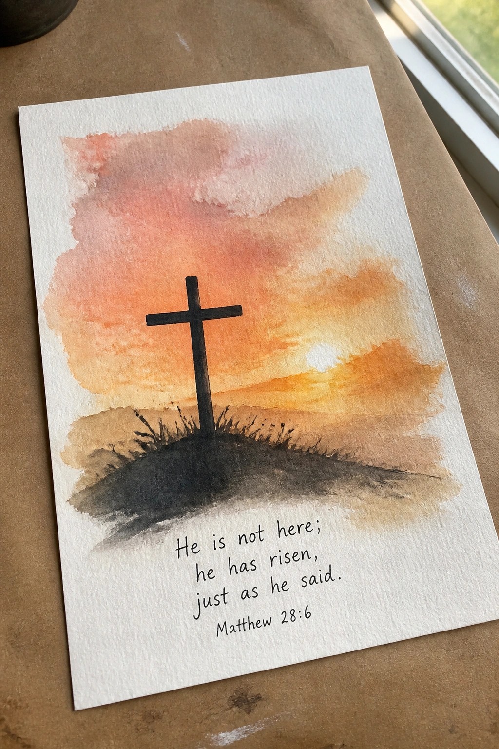 Watercolor of black cross on hill at orange-pink sunset with Matthew 28:6 verse below