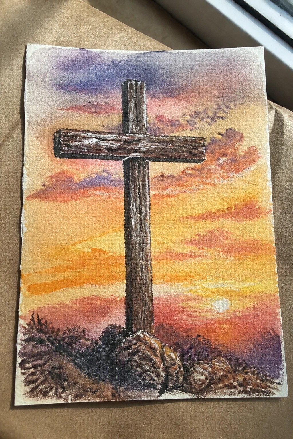 A painting of a brown wooden cross standing upright on rocks with a colorful sunset sky and clouds behind it.