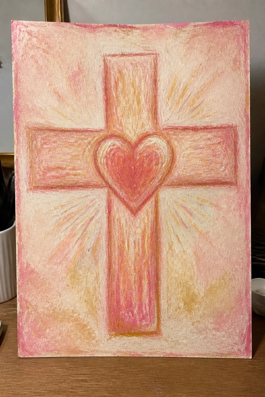 Pink cross with central heart and radiating yellow-orange rays on textured background.