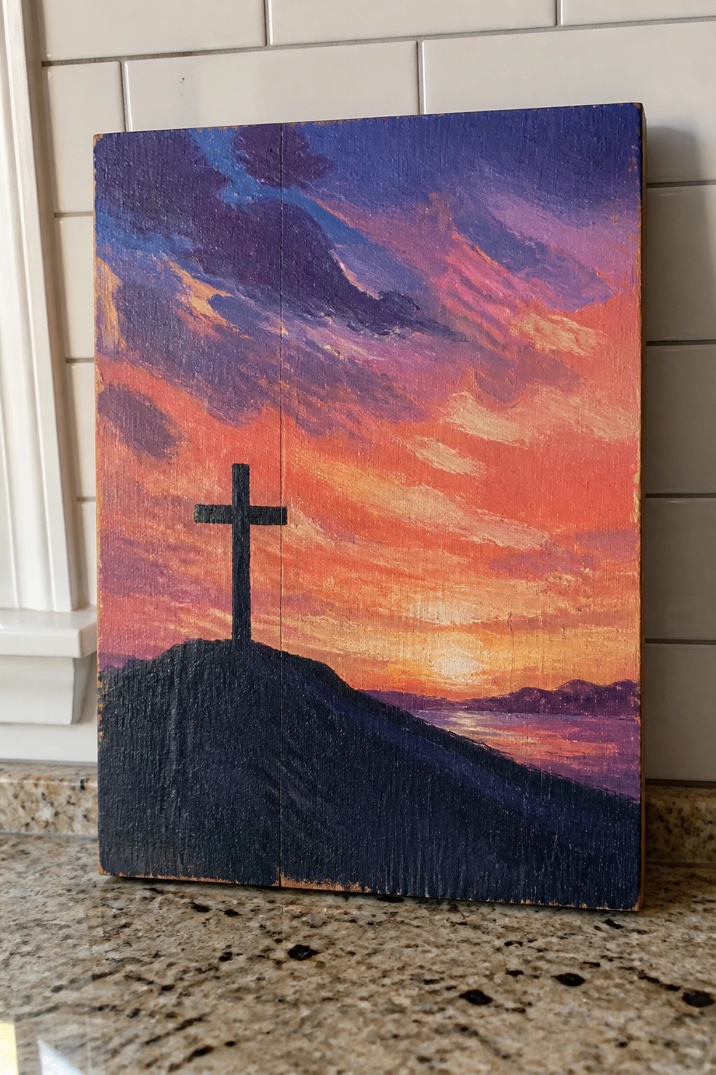 Vibrant sunset painting on wood with black cross silhouetted on dark hill above water.