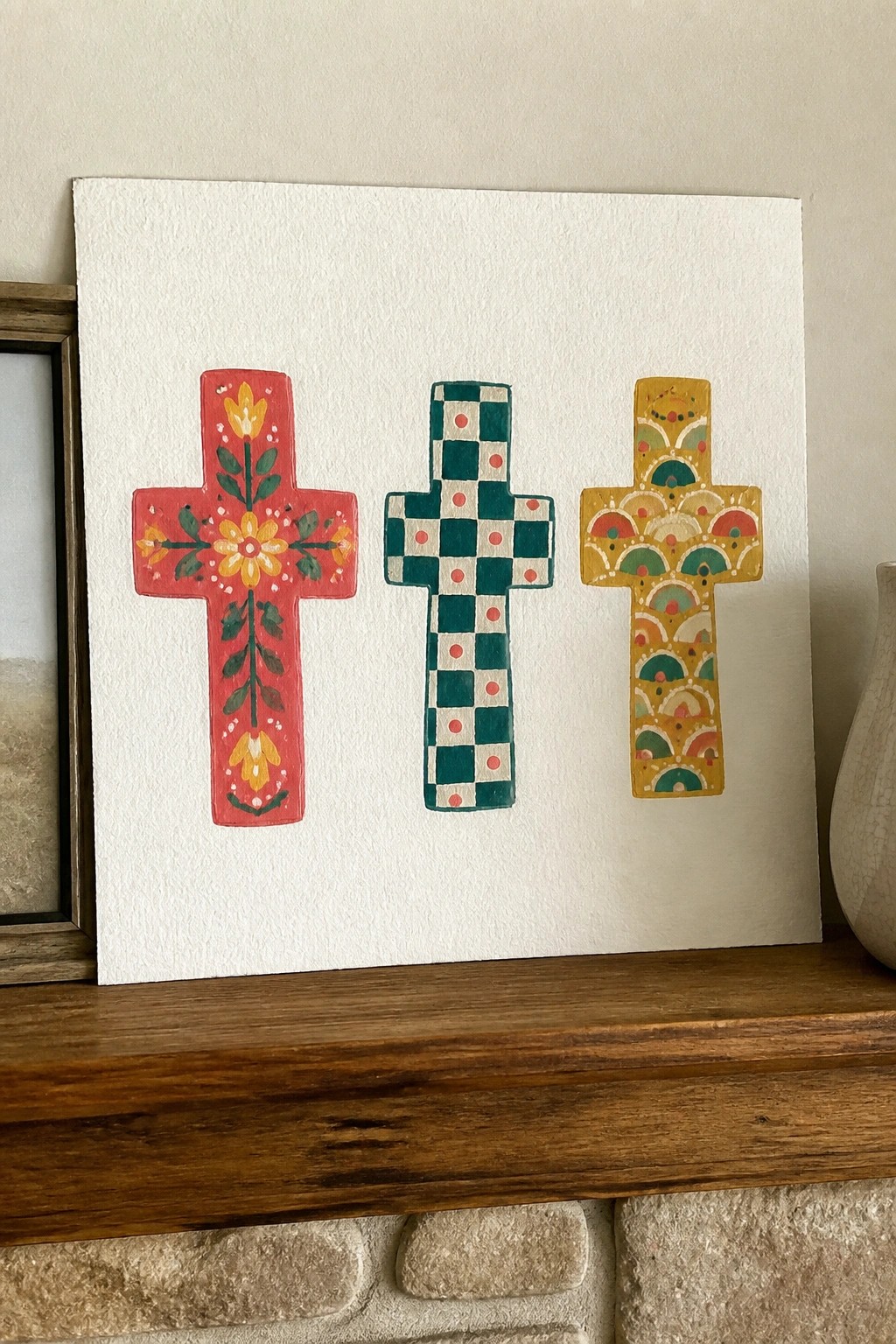 Three vibrant patterned crosses on white paper resting on wooden mantelpiece