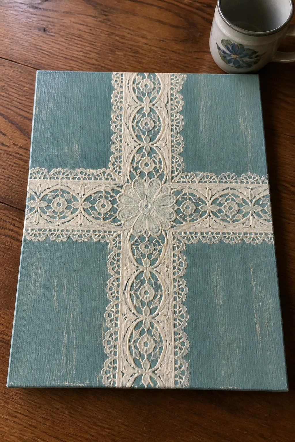 Teal canvas with raised white lace cross on wooden table beside floral mug