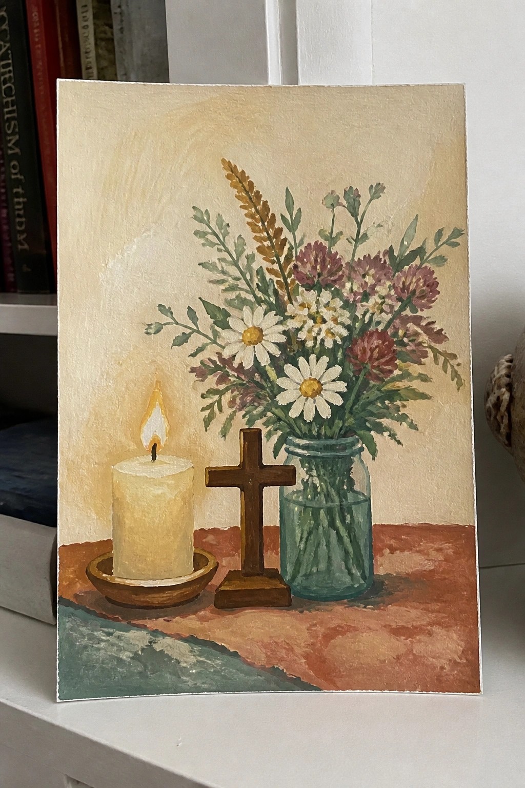 A painting shows a lit candle in a dish, a wooden cross, and a glass jar of daisies and wildflowers on a reddish surface.