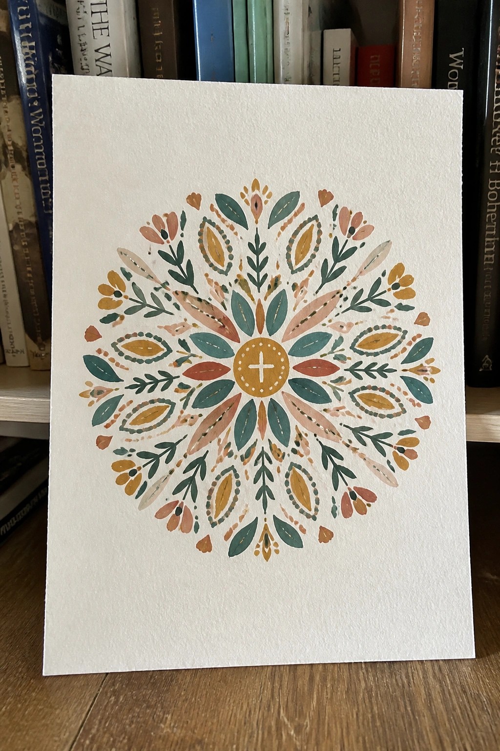 Watercolor floral mandala with teal, orange, and yellow patterns on white paper before books