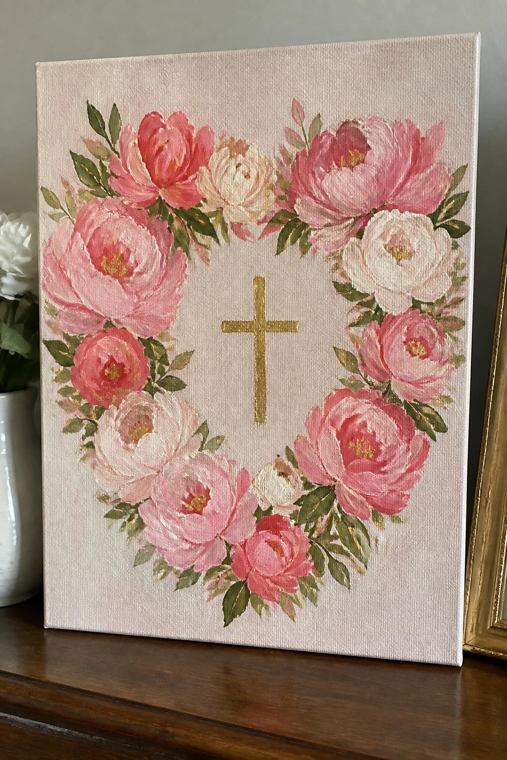 A canvas with a wreath of pink and white flowers around a gold cross on a light background.