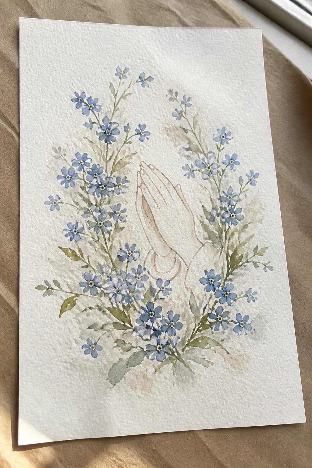 Watercolor of praying hands amid blue forget-me-not flowers and soft green leaves.