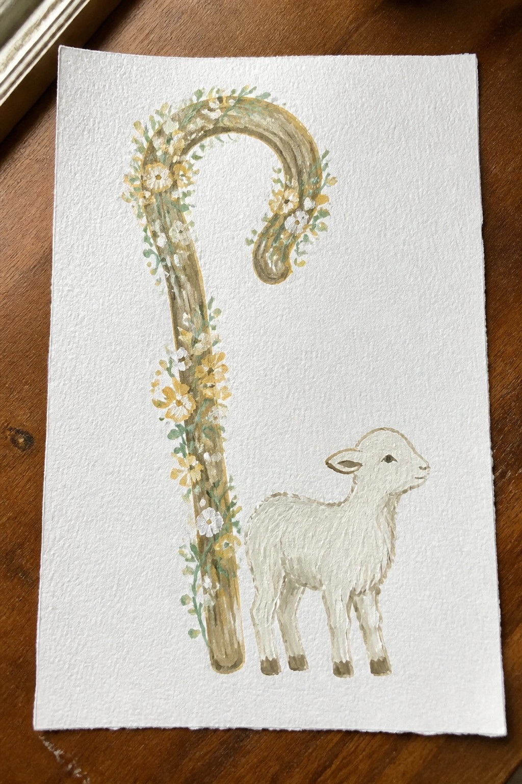 Watercolor of a floral shepherd's crook beside a small white lamb on paper