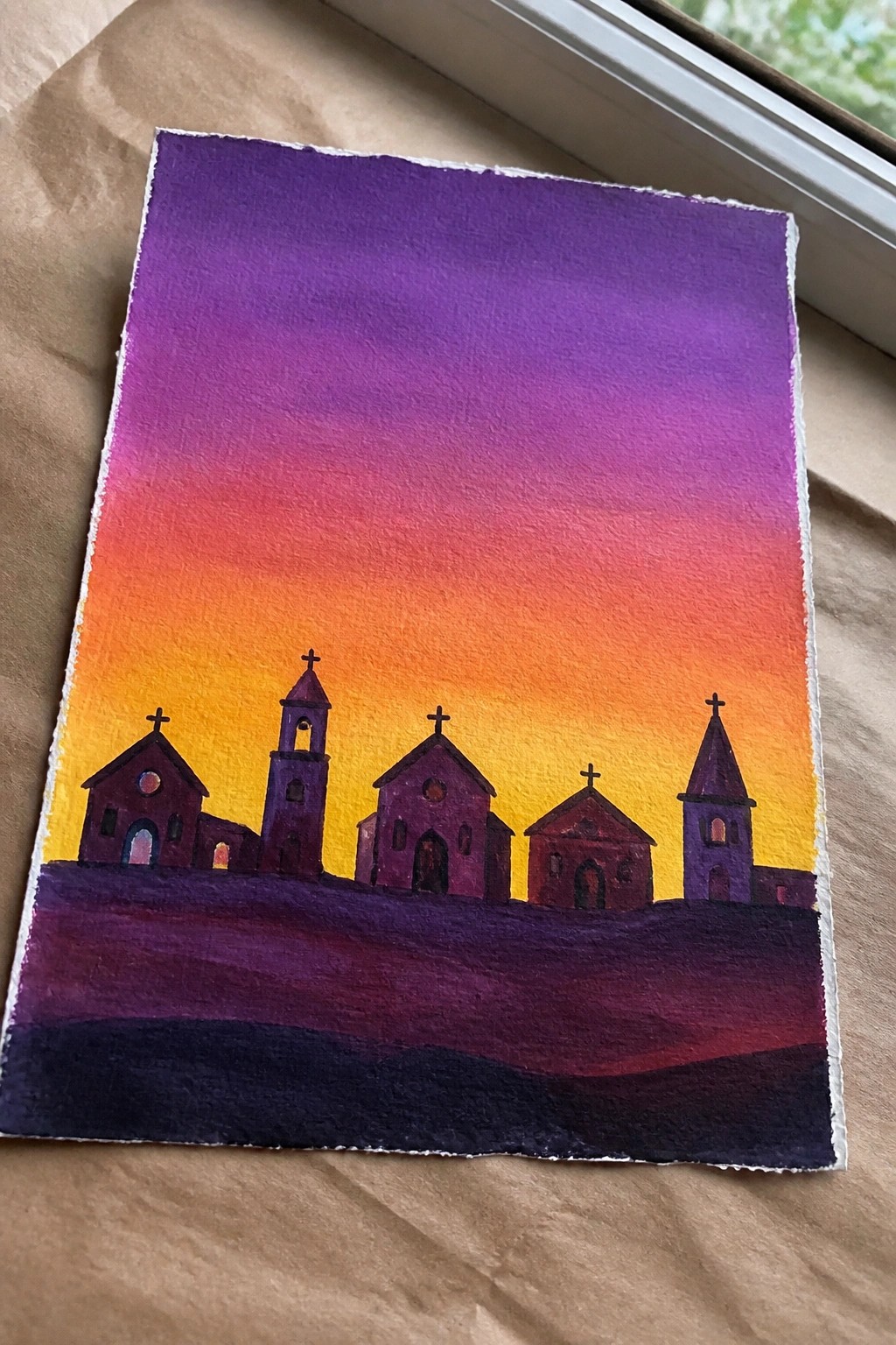 Watercolor painting of purple church silhouettes with crosses under a vibrant orange-purple sunset.
