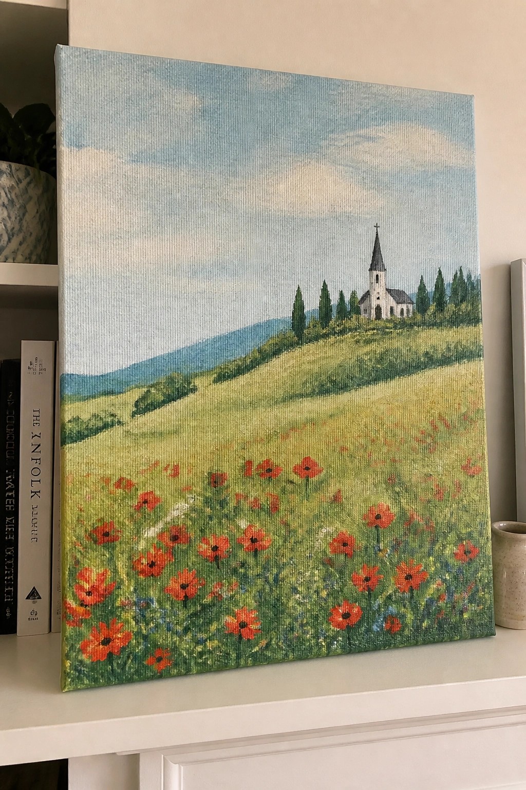 Painting of red poppies in meadow with church on hill, displayed on shelf with books