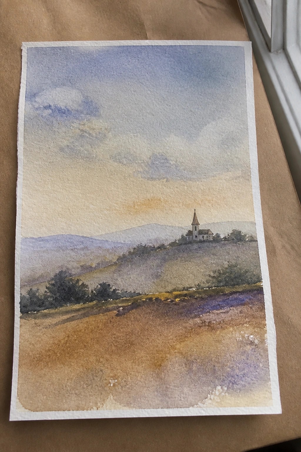 Watercolor landscape of church on hillside with rolling fields, distant mountains, and cloudy sky.
