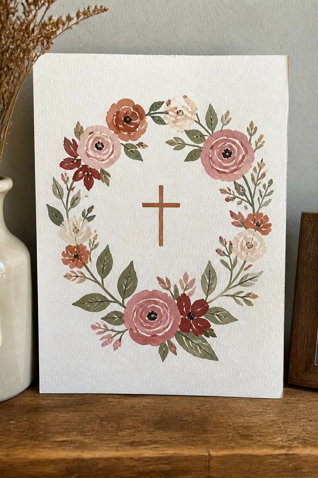Watercolor wreath of pink flowers surrounding brown cross on wooden surface
