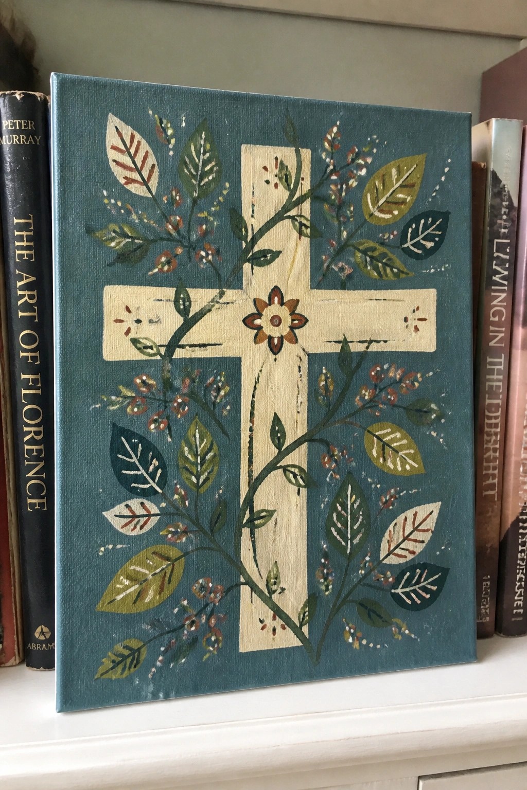 Floral cross painting on teal background beside books on white shelf