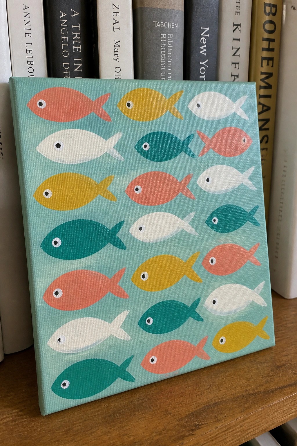 Colorful fish painting on teal canvas placed before a row of books.