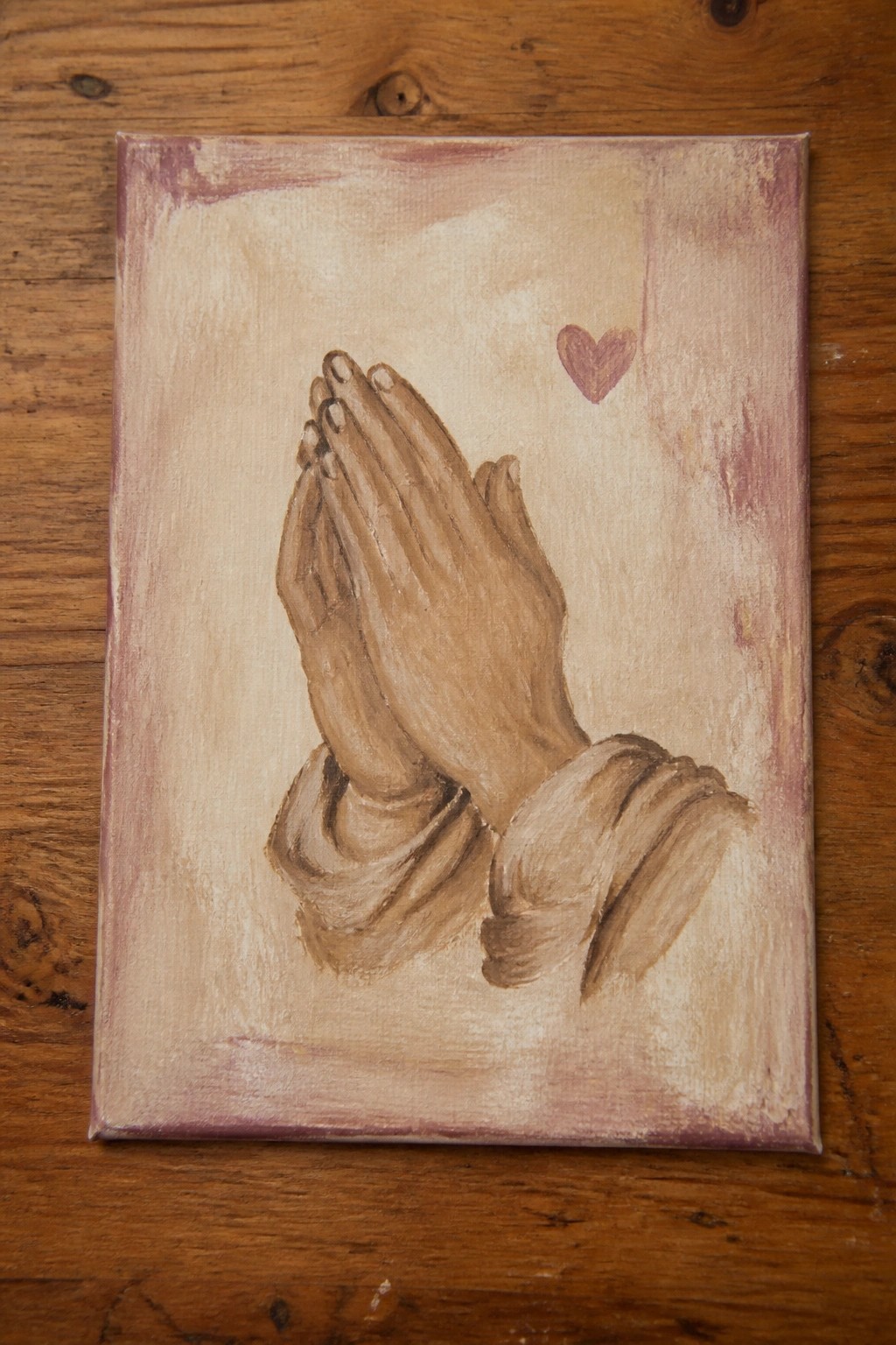 Painting of praying hands with small pink heart on textured canvas
