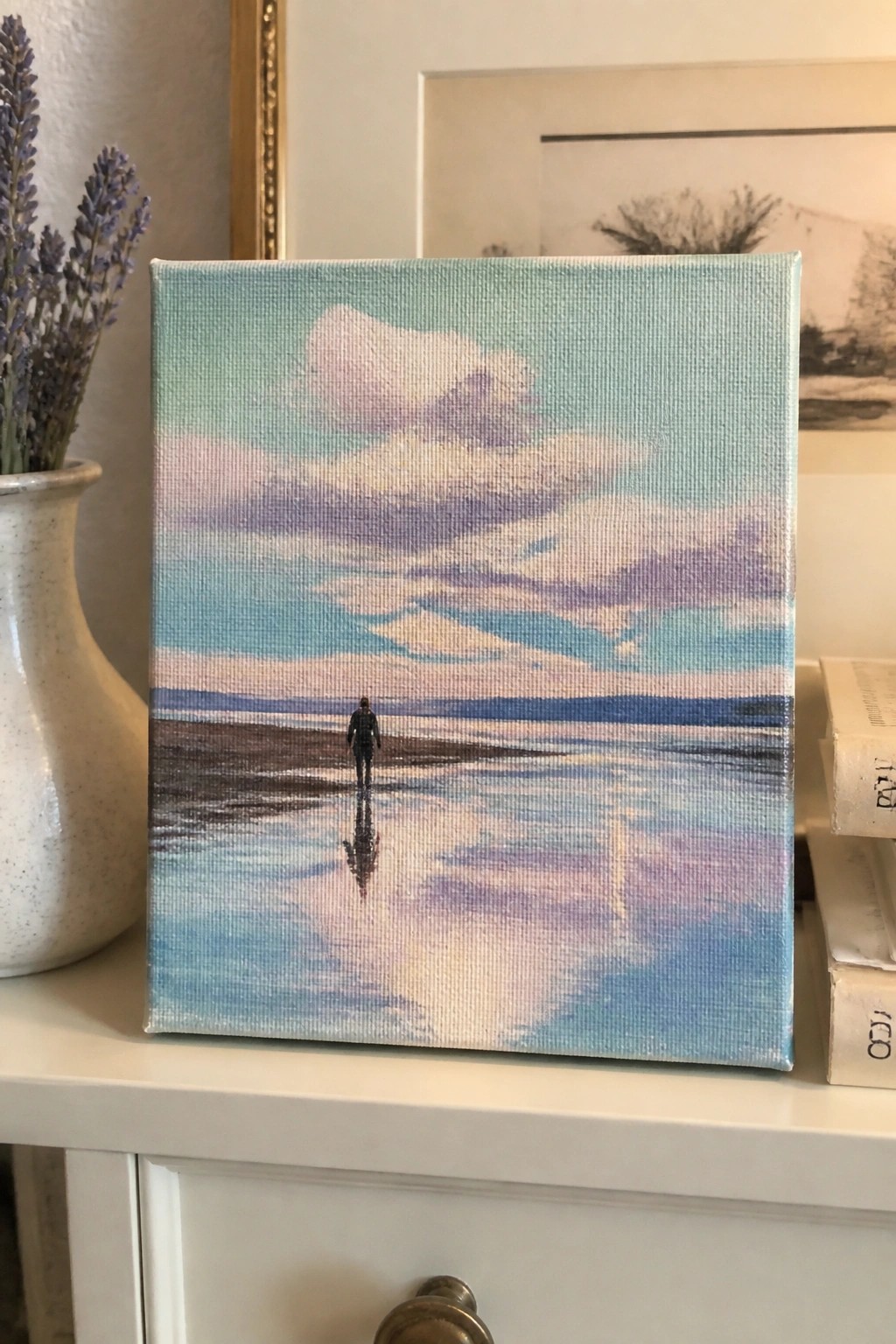 Small canvas beach painting with lone figure and sky reflections beside lavender vase and books.