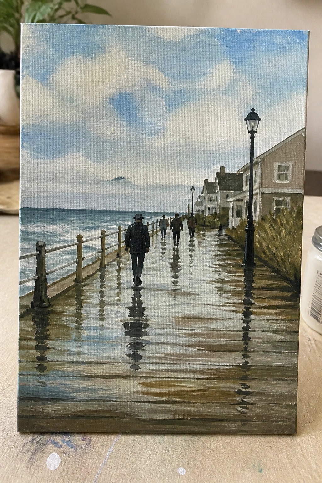 Rain-soaked wooden boardwalk by the sea with walking figures and distant houses.