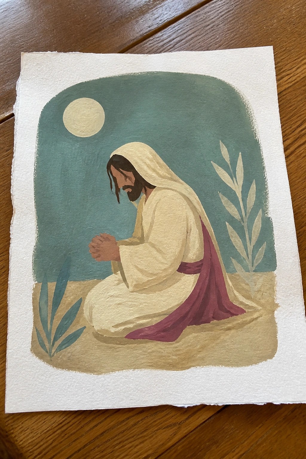 Watercolor of Jesus in white robe with maroon sash kneeling in prayer under moon at night