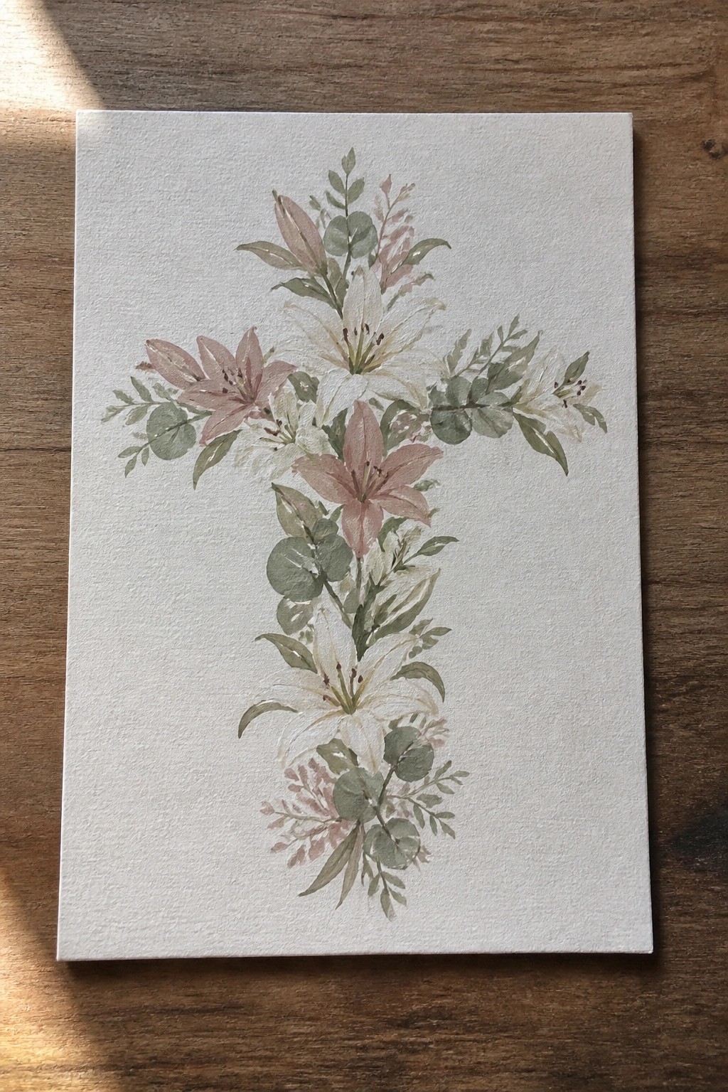 Watercolor cross of pink and white lilies with green leaves on paper over wood