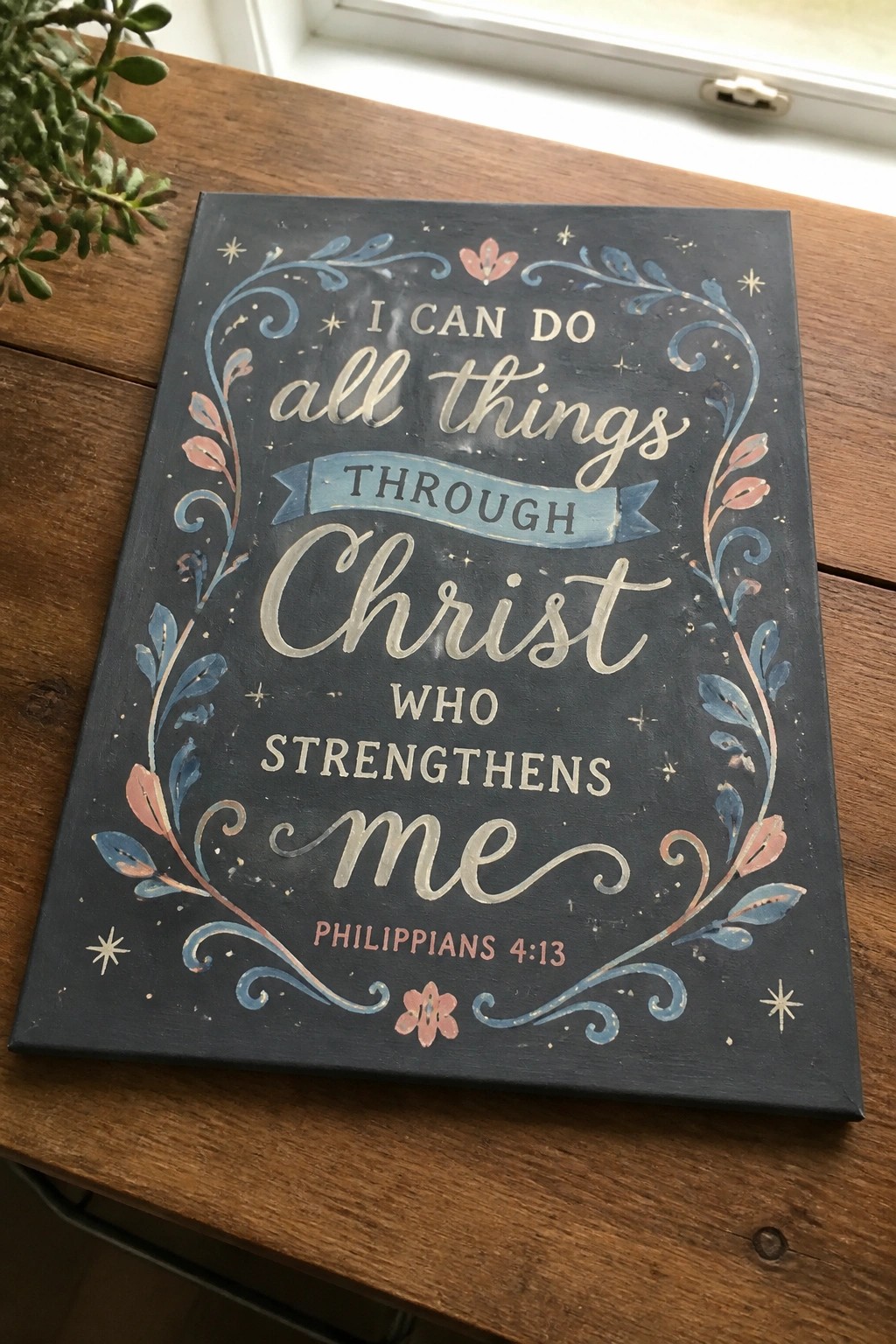 Dark blue sign with pink-blue floral border and Philippians 4:13 verse on wooden table.