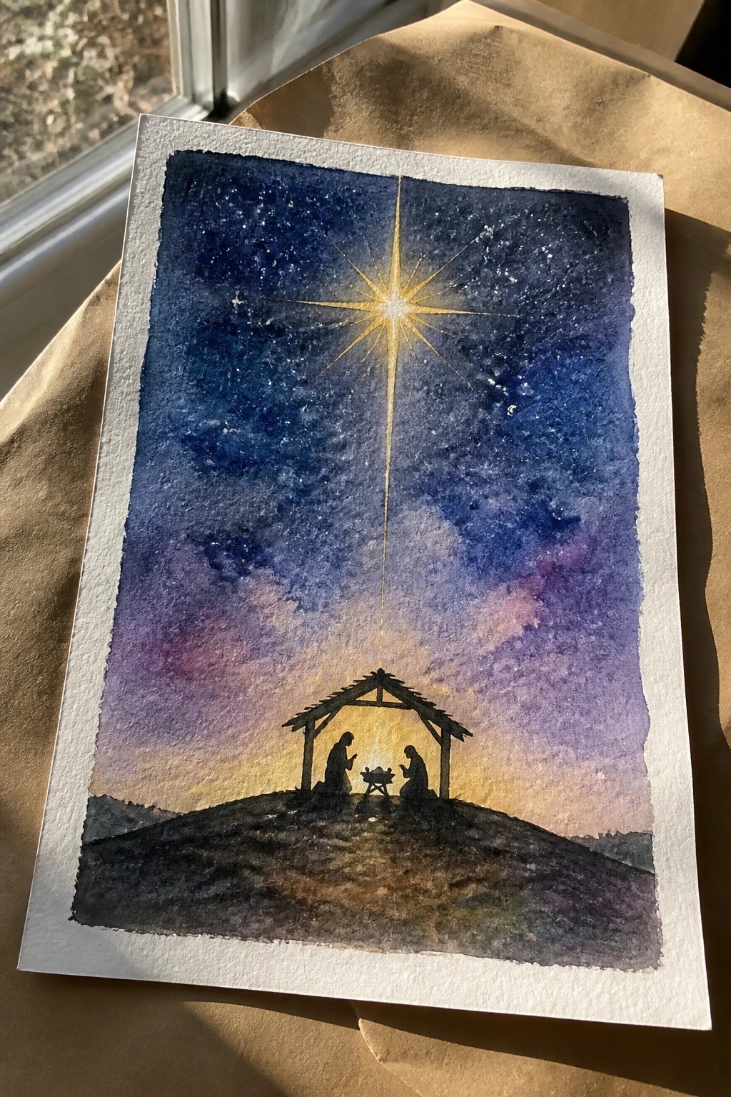 Watercolor nativity silhouette with two kneeling figures under bright star in purple-blue sky