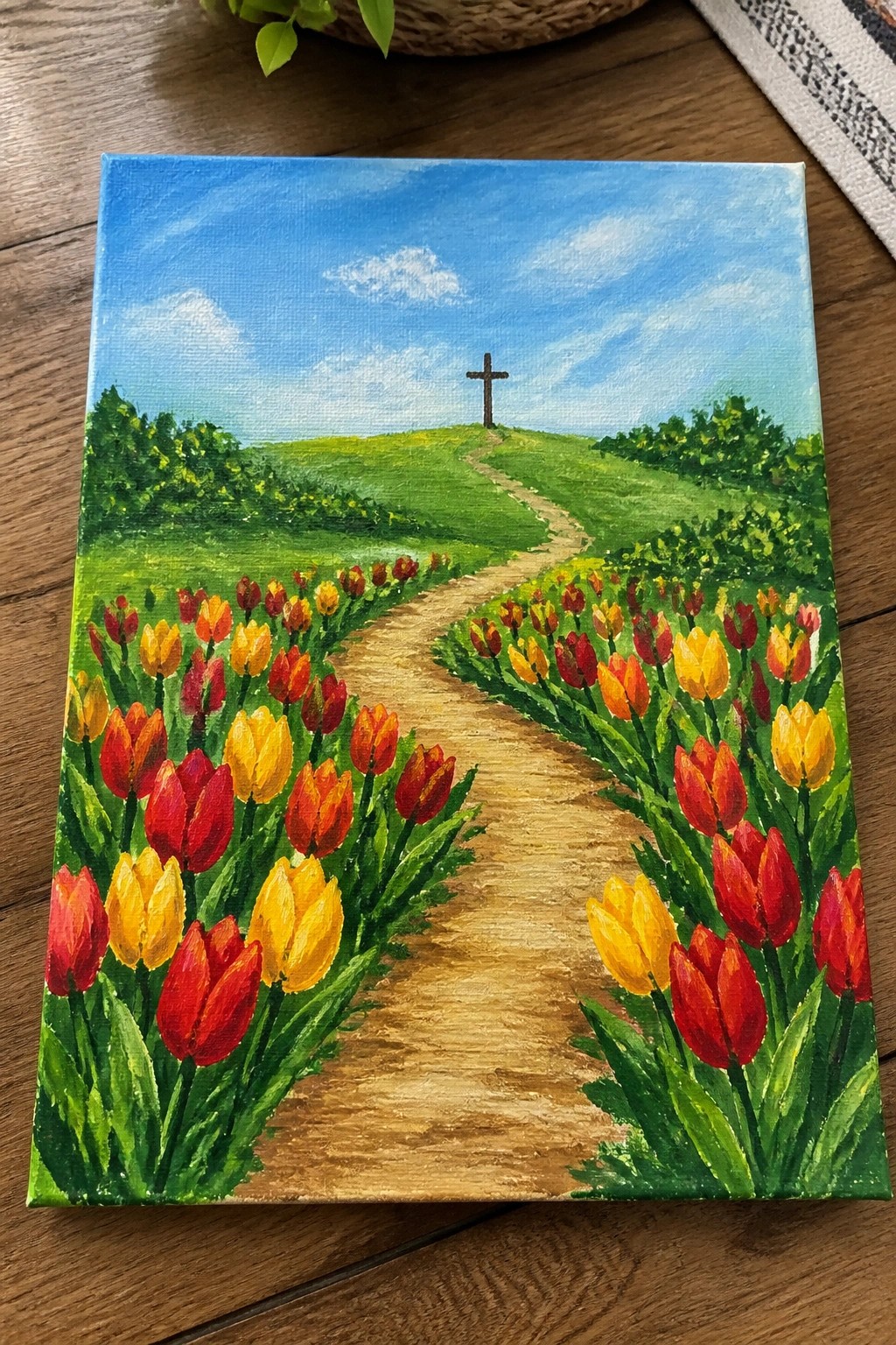 Vibrant painting of tulips lining a winding path to a hilltop cross under blue skies