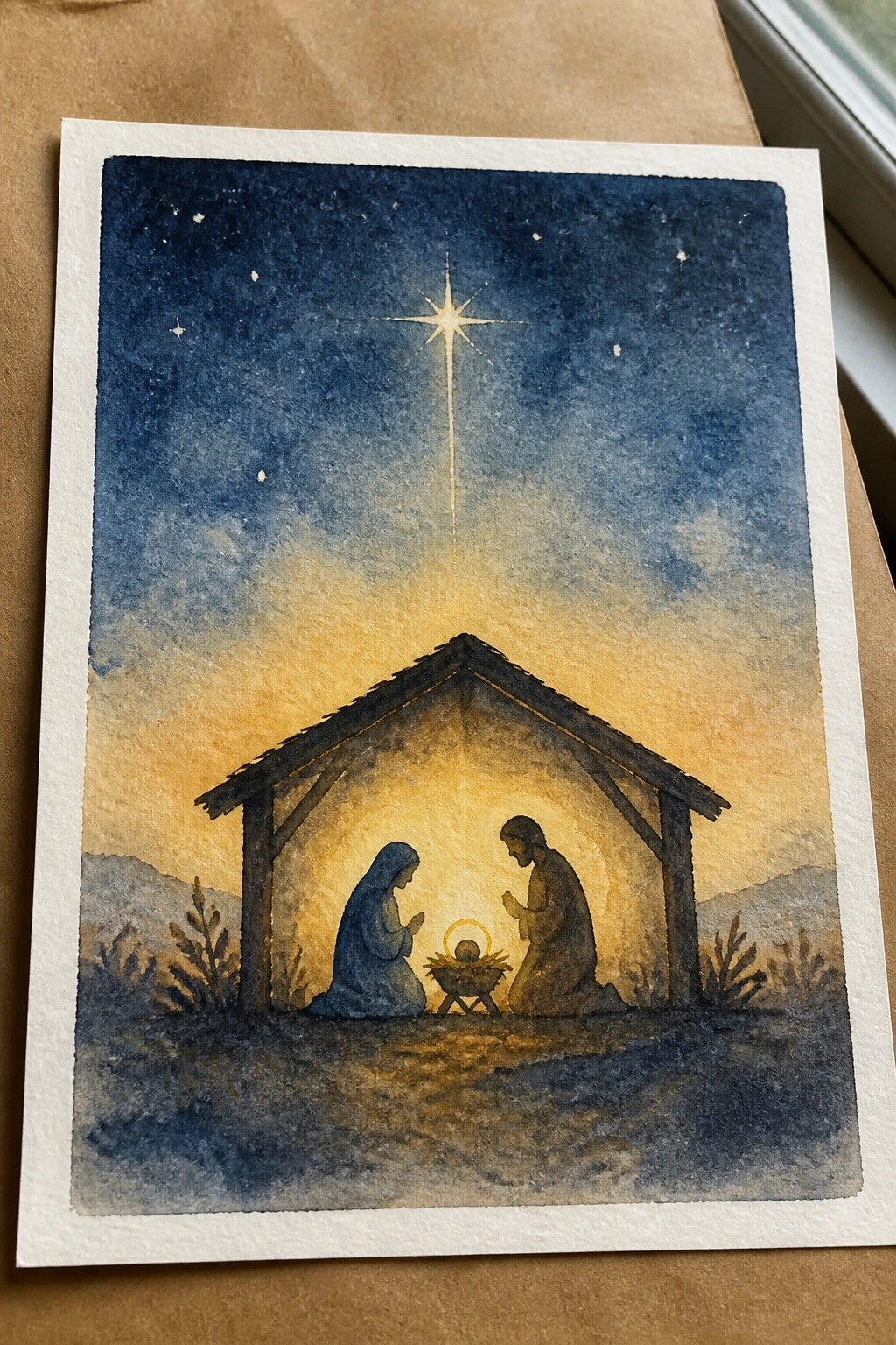 Watercolor nativity with Mary in blue kneeling by Joseph and baby Jesus under bright star