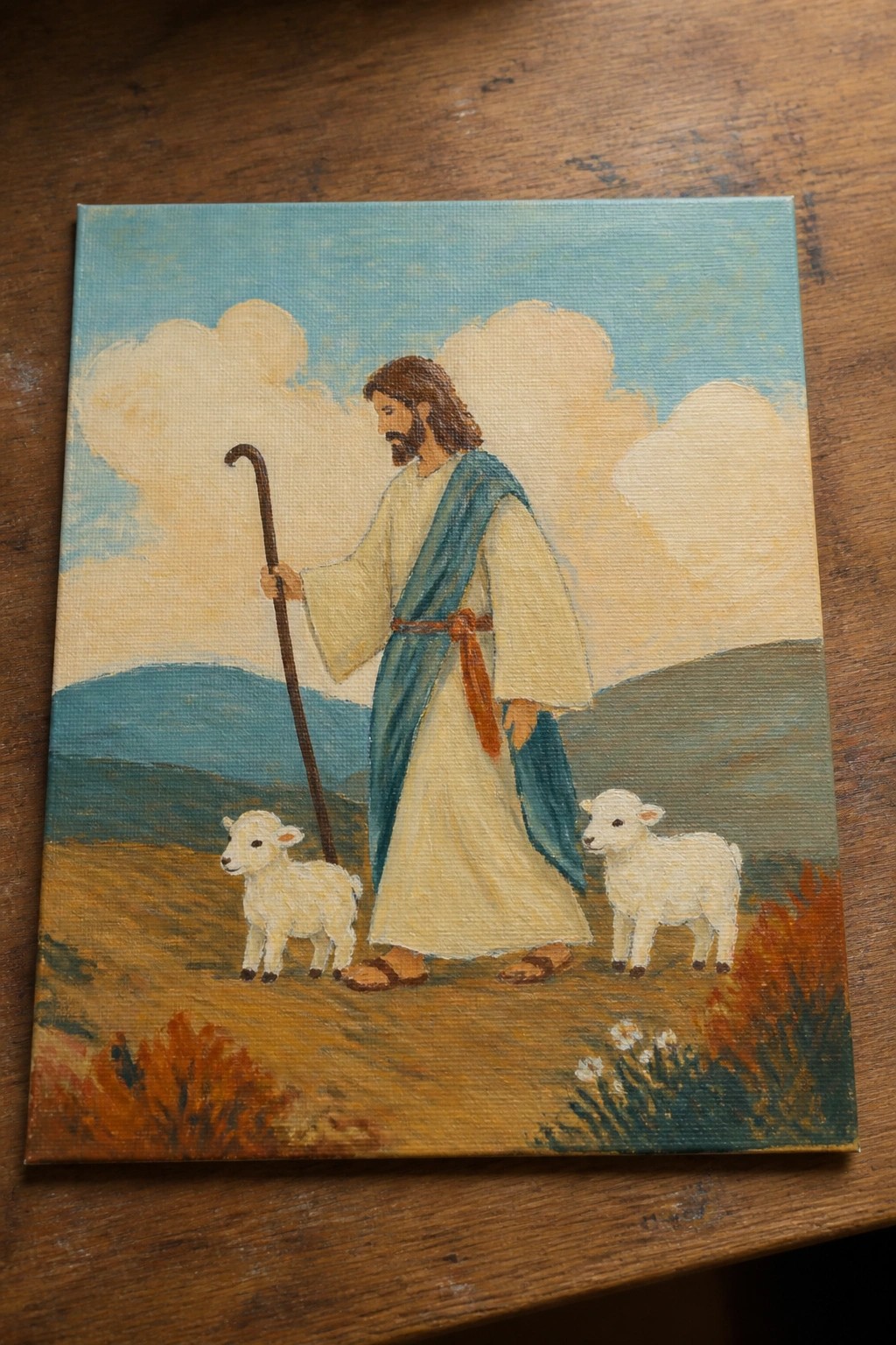 Bearded shepherd in white robe with blue sash holds staff beside two white lambs in landscape.