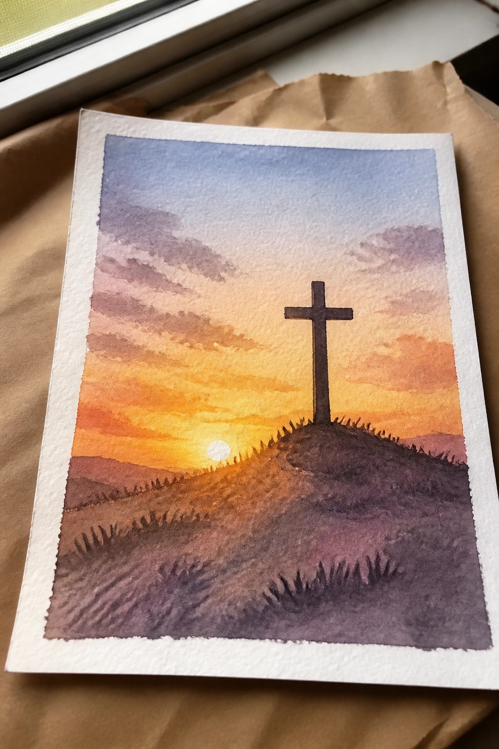 Watercolor sunset with a silhouetted cross on a hill under orange and purple skies.